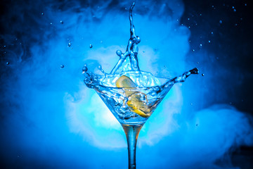 Martini cocktail glass in hand splashing on dark toned smoky background or colorful cocktail in glass with splashes and olives. Party club entertainment. Mixed light. Selective focus