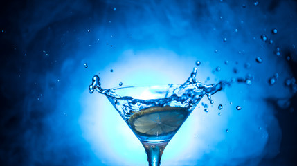 Martini cocktail glass in hand splashing on dark toned smoky background or colorful cocktail in glass with splashes and olives. Party club entertainment. Mixed light. Selective focus