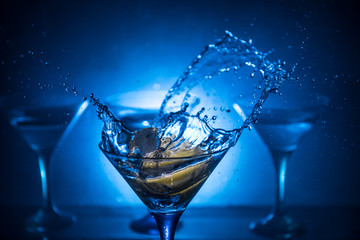 Martini cocktail glass in hand splashing on dark toned smoky background or colorful cocktail in glass with splashes and olives. Party club entertainment. Mixed light. Selective focus