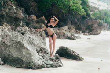 Sexy tanned brunette girl in black swimwear sunbathes on beach with rocks.
