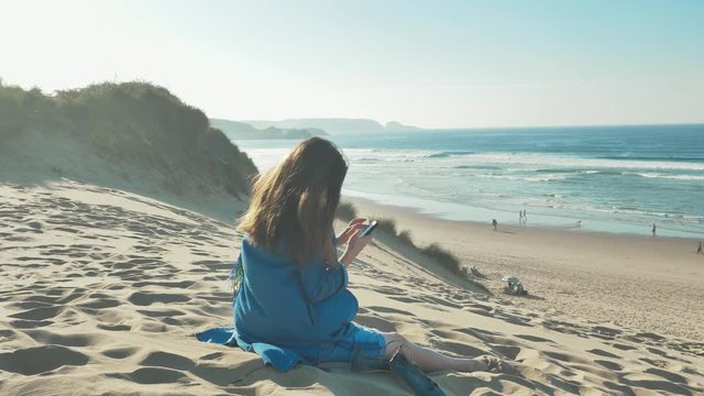 A fully clothed young woman on a sandy beach in a sunny day, sitting on the sand, taking a picture of the relaxing panorama with her smartphone.