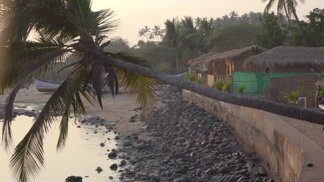 Panning shot across row of grass thatched colourful beach huts on the shoreline of Indian, Goa beach. View of coastal waters in foreground.
