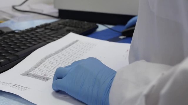 Doctor or a scientist is working on his research on the computer, cross checking he data on a paper sheet.