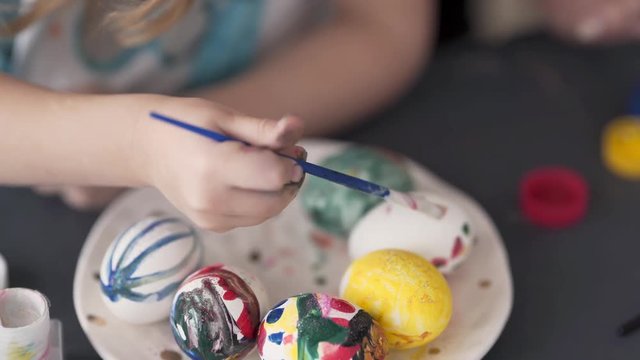 A close up video of the egg painting process. A little girl paints a ceramic egg. She uses a brush in order to make unequal dots or red colour. The background is blurred.