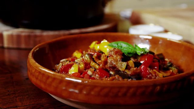 Video zooming in on cooked ratatouille recipe in a rustic ceramic dish sitting on a wooden country table.