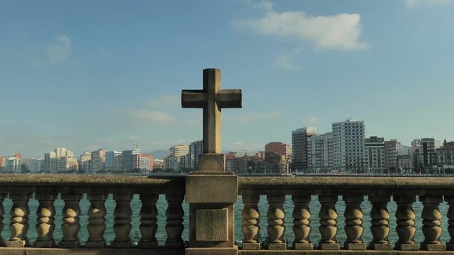 A stone cross facing the sea in Gijon (Asturias, Spain), in front of tall buildings. Shot taken in August 2018.