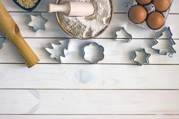 Cutter for cookies and ingredients in one line. Light wooden background with a bowl of flour, eggs, butter and cookie cutters for ginger Christmas tree cookies, star, bell. Save the space. Background 