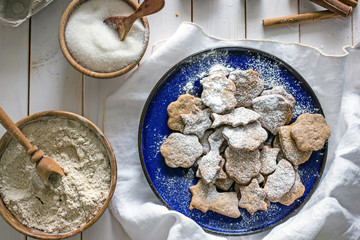 Shortbread homemade cookies of different shapes in a blue plate and adjusted with icing sugar. Next to the ingredients for baking, white wooden background, top view.