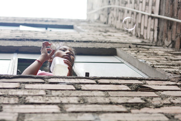 Native american teenager girl blowing soap bubbles from the window of brick building.