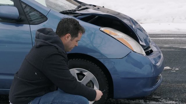 Man Squats Near Wrecked Car Upset And Contemplates What To Do Next. Camera is fix on man crouching near the front passenger side of his wrecked car. Man seems to be thinking while rubbing his head.