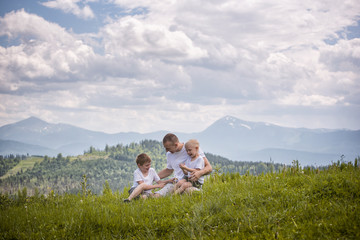 Happy father with his two young sons sitting on the grass on a background of green forest, mountains and sky with clouds. Friendship concept.