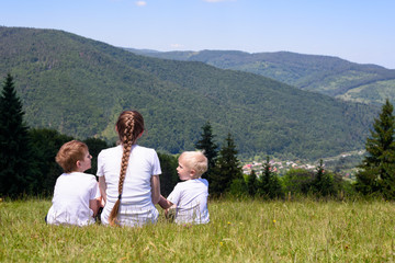 Mother and two young sons sitting on grass on the background of coniferous forest and mountains