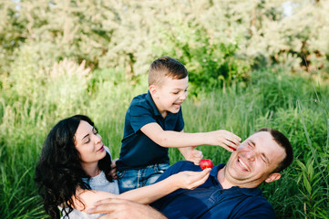 Happy young family on vacation eating strawberries together, outdoors. The father, mother, little boy having picnic on blanket, grass, nature. Portrait of mom, dad, son. The concept of family holiday.