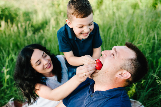 Happy young family on vacation eating strawberries together, outdoors. The father, mother, little boy having picnic on blanket, grass, nature. Portrait of mom, dad, son. The concept of family holiday.