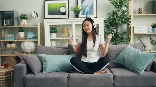 Beautiful Asian girl is listening to music through headphones and dancing moving hands and body sitting on sofa at home alone. Youth culture and gadgets concept.
