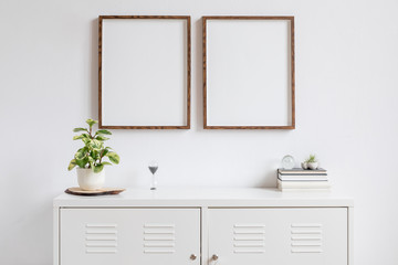 Fototapeta na wymiar Minimalistic home decor of interior with two brown wooden mock up photo frames above the white shelf with books, beautiful plant in stylish pot and home accessories. White wall. Concept of mockup.