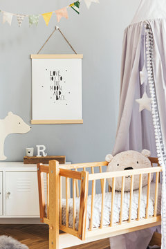 Stylish and cute scandinavian decor of newborn baby room with mock up poster , white design furnitures, natural toys, hanging grey canopy with wooden cradle, pillows, accessories and teddy bears. 