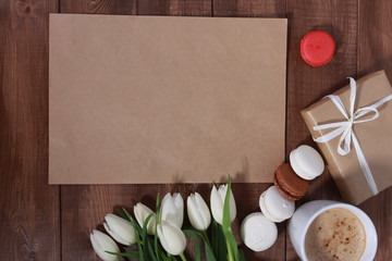 Bunch of white tulips, gifts,  macaroons, cup of coffee on brown  wooden table. Greeting concept. Spring, women or mothers day, cakes, cappuccino, blank sheet of paper