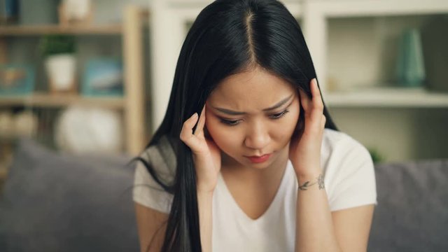 Unhappy Asian female student is feeling bad having headache and trying to release pain massaging her head touching temples with sad face. Sick youth concept.