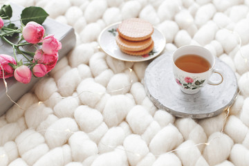 Cup of tea, cookies, pink rose flowers and book on white knitted woolen merino chunky blanket. Breakfast in bed. Cozy spring homely scene. Home decor.