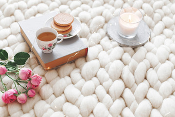 Fototapeta na wymiar Cup of tea, cookies, pink rose flowers, book and lighted candle on white knitted woolen merino chunky blanket. Breakfast in bed. Cozy spring homely scene. Home decor.