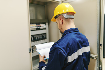 Electrician in protective suit and helmet holding clipboard and trying to fix glitch while standing next to dashboard in heavy industry plant.