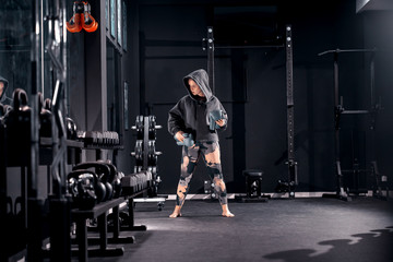 Fototapeta premium Young muscular Caucasian woman with hoodie on head holding boxer gloves and standing in gym barefoot. Night workout concept.