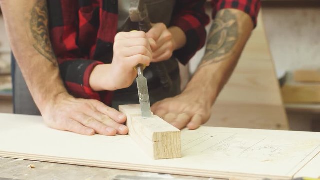 Hardworking little caucasian boy in safety glasses and an apron works with chisel in a carpentry shop being supervised by adult professional woodworker.