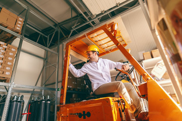 Fototapeta na wymiar Smiling Caucasian worker in white uniform and protective helmet on head driving forklift in warehouse.