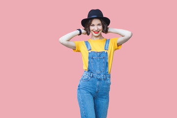Portrait of happy young woman in yellow t-shirt, blue denim overalls with makeup and black hat...
