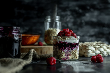 view on a dark, wooden table top, on which stands in a glass jar fresh yogurt with cereal, pits and raspberries, and ingredients for making it