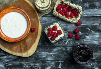 top view on a dark, wooden table top, on which stands in a glass jar fresh yogurt with cereal, pits and raspberries, and ingredients for making it