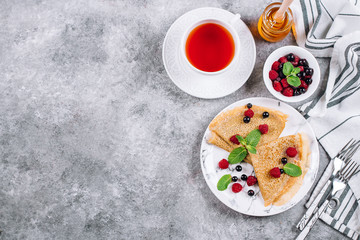 Obraz na płótnie Canvas Delicious Crepes Breakfast on gray concrete table background. Orthodox holiday Maslenitsa. Pancakes with berry black currant, raspberry, jar of honey and mint. White cup of tea. Top view, copy space