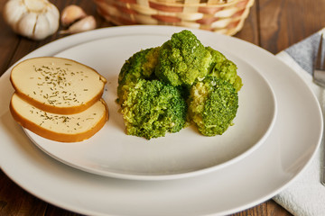 Dish of broccoli and two slices of defatted cheese next to a napkin, fork, basket with bread and garlic on a dark wooden background. Close-up