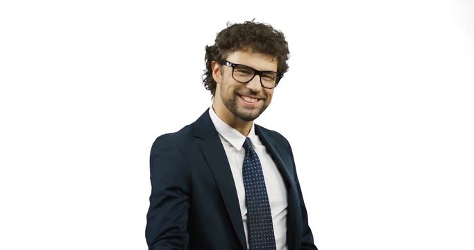 Young attractive Caucasian businessman in glasses, suit and tie standing in front of the camera and doing yes, well done gesture with a hand. White wall screen.