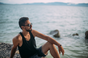 A young man sits on the beach. Handsome man in sunglasses looks at the horizon on the ocean at sunset.