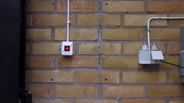 Pan right from a black steel door to a yellow and orange brick and mortar wall, where a light switch, fusebox and electricity outlet are mounted