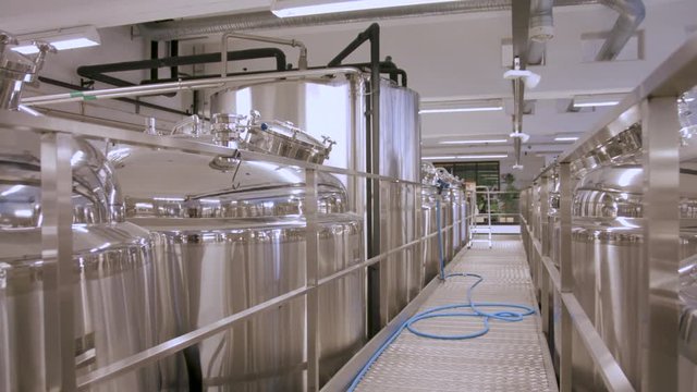 Slow pan right of a steel walkway between rows of stainless steel fermentation tanks in a brewery
