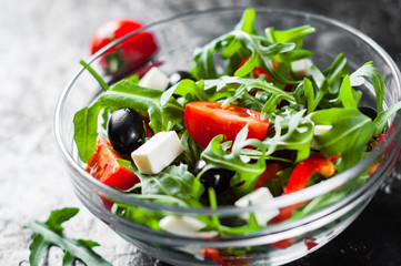 fresh green salad with arugula, tomatoes, cheese, pepper and olive in glass bowl on dark background