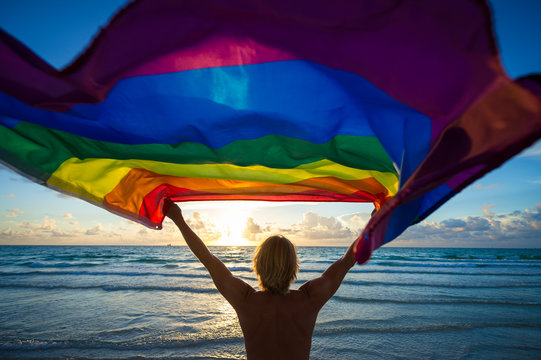 Colorful silhouette of a man with blond hair holding a gay pride rainbow flag blowing in the wind on a tropical beach with golden sun