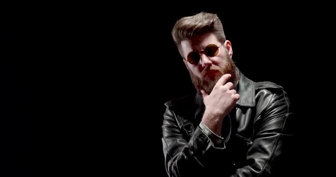 Serious man with stylish beard on black backgroung. Man in black leather clothes with stylish haircut and nice big beard.