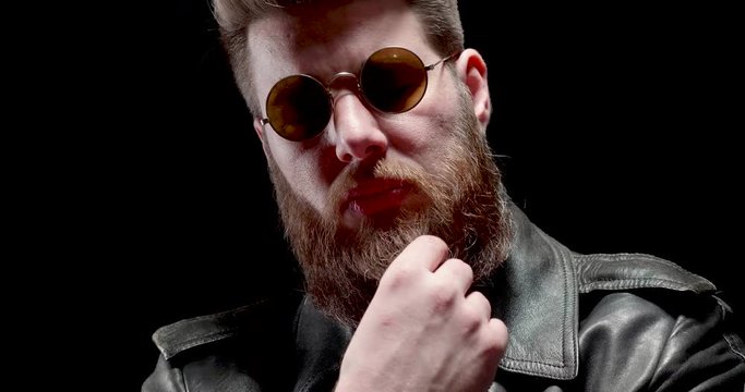 Serious man with heavy metal look chews tabacco and touches his beard. Biker on a black background.