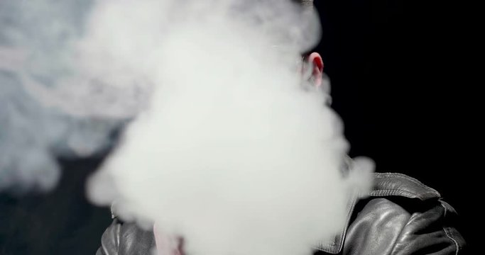 Heavy metal stylish guy with big beard vaping. Exhales a lot of smoke that covers the whole picture. White smoke on black background.