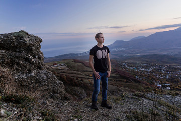 Naklejka na ściany i meble Portrait in growth.. Young attractive man standing on the top of mountains looks into the distance at Violet sunset. 