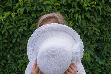 Little girl covers her face with a white hat on a green grape leaf background