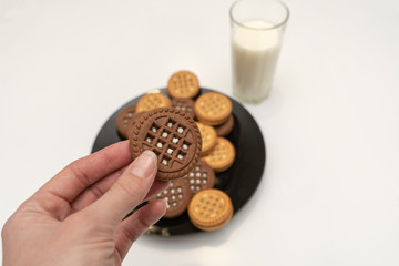 Close-up of chocolate chip cookie in hand. Big black plate full of cookies and a glass of milk on white table