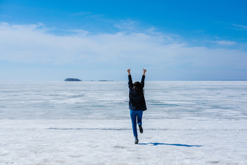 The girl jumps up and holds his hands up with a thumbs up finger, fun and enjoying life on the background of the icy sea and blue sky.