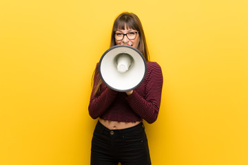 Woman with glasses over yellow wall shouting through a megaphone