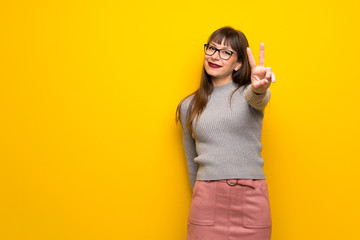 Woman with glasses over yellow wall smiling and showing victory sign