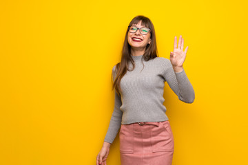 Woman with glasses over yellow wall saluting with hand with happy expression